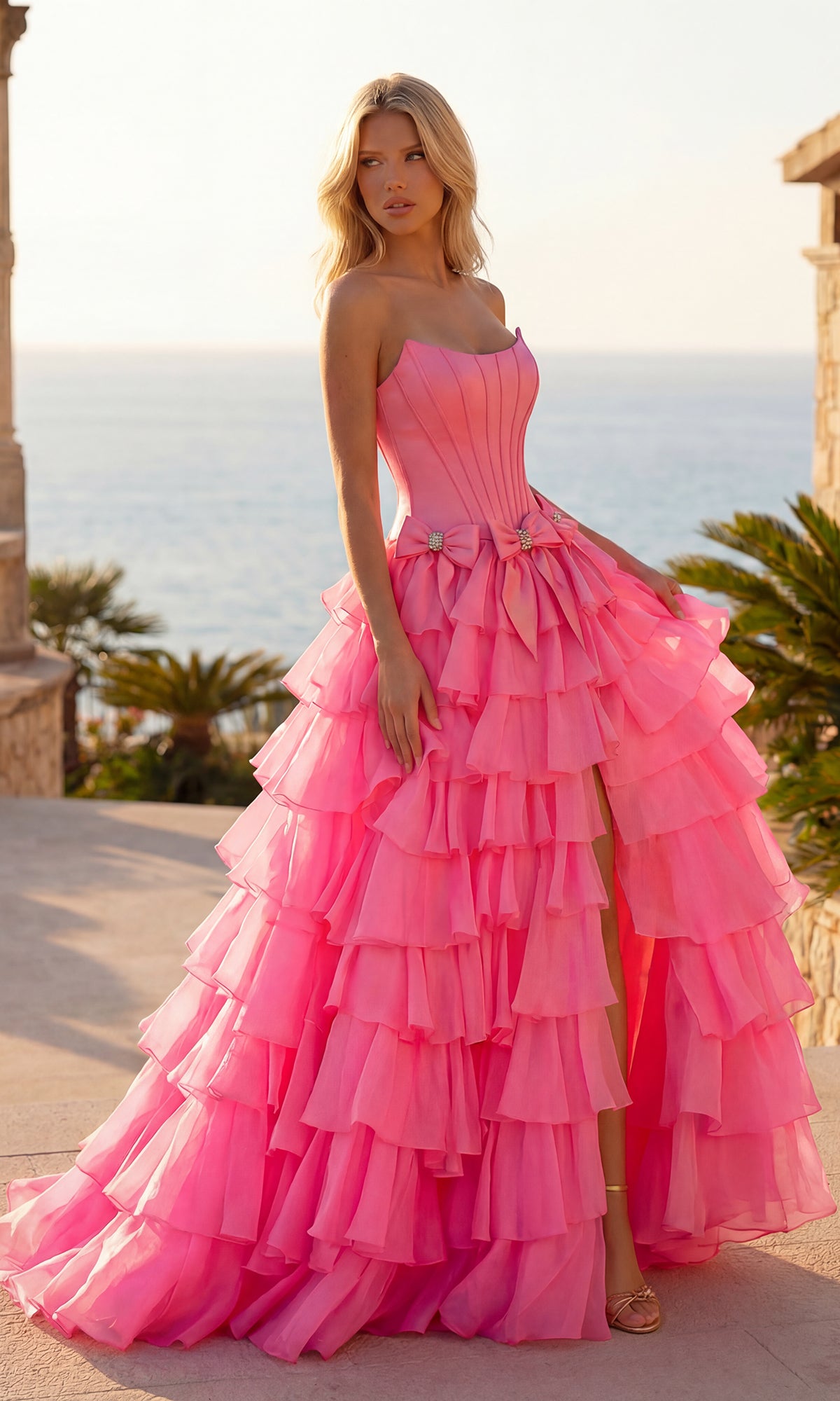 A woman stands outdoors in front of palm trees and the sea, wearing the bright pink PromGirl 250071 tiered prom ball gown with bows.